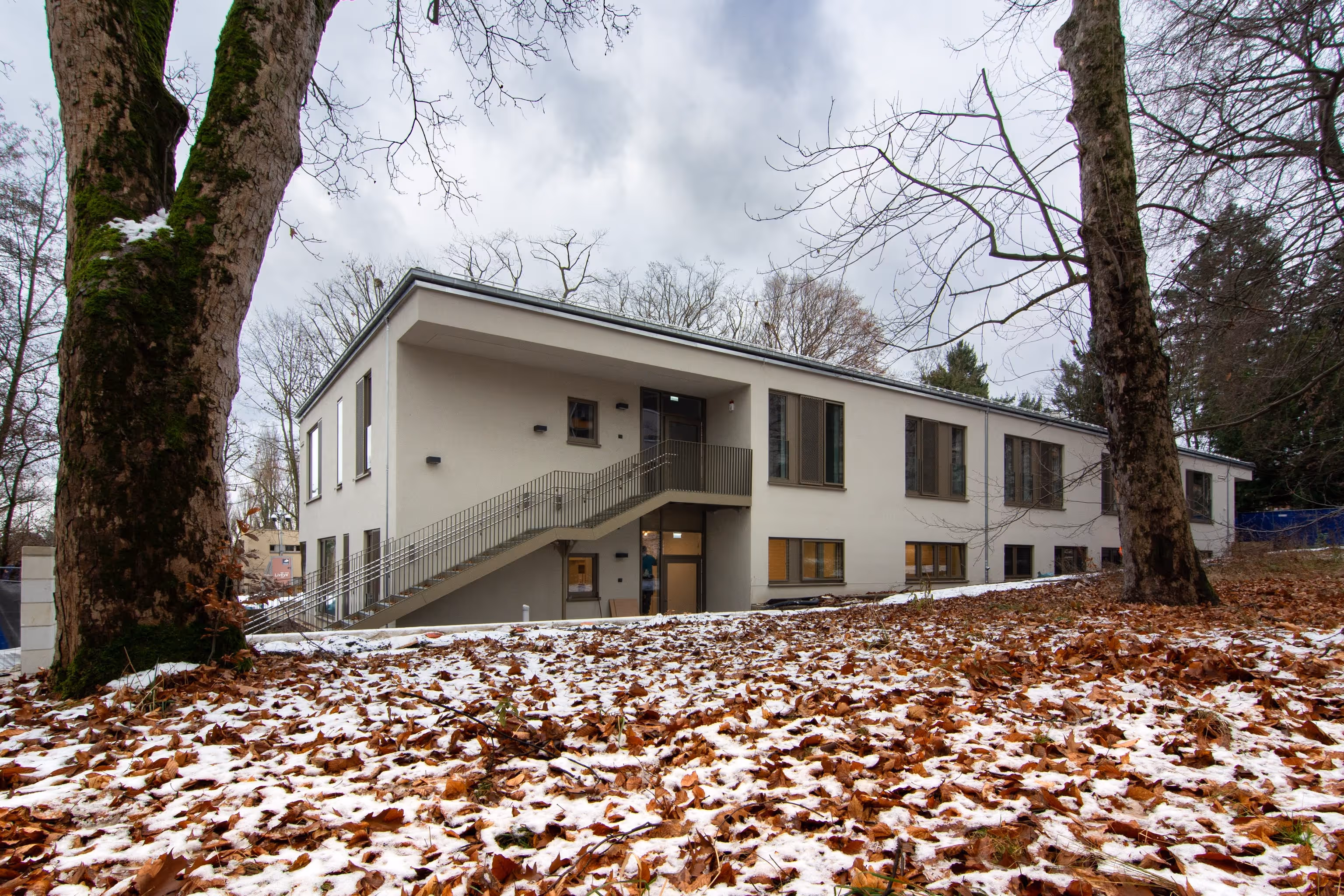 Modern flaches Kita-Gebäude mit Außentreppe, Herbstlaub und leichtem Schneefall zwischen Bäumen.