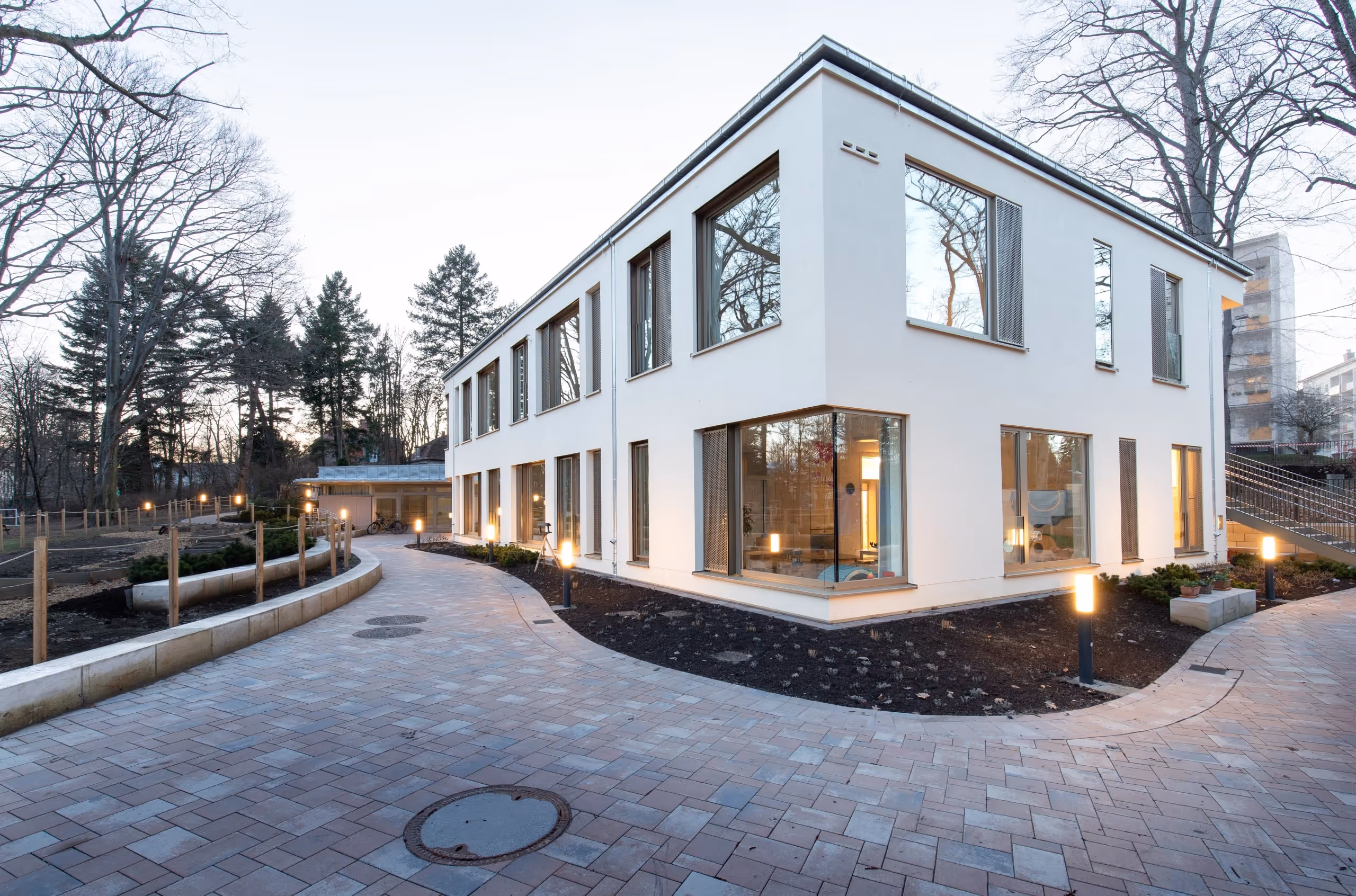 Modern zweistöckiges Kita-Gebäude mit großen Fenstern, beleuchteten Wegen und kahlen Bäumen im Hintergrund.