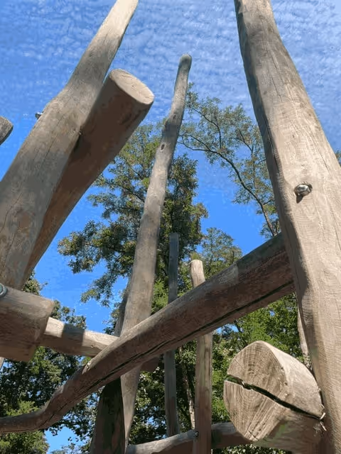 Blick von unten durch ein Klettergerüst aus Holzbalken in Baumkronen und blauen Himmel.