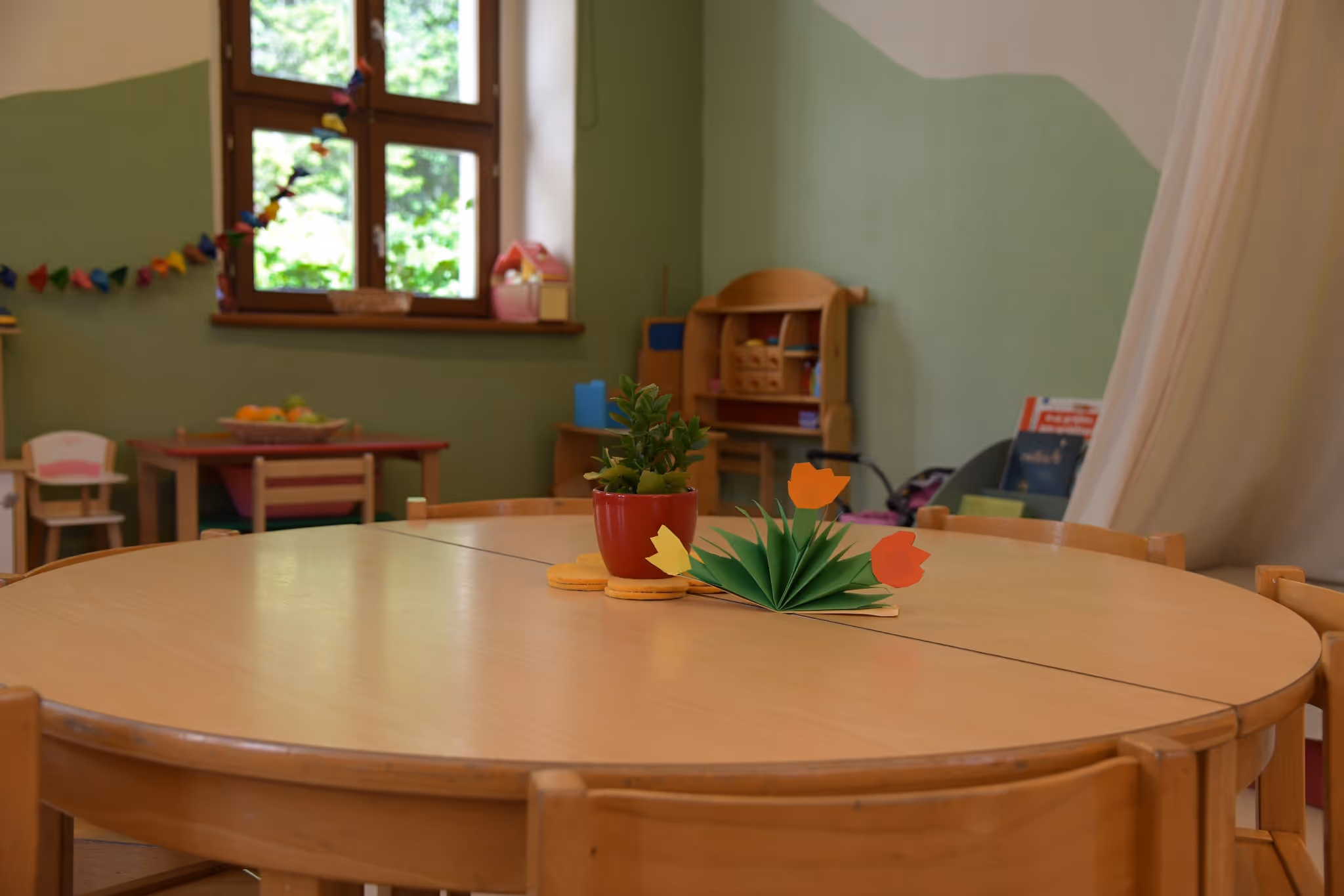 Runder Holztisch mit Papierblumen als Tischschmuck in einem Gruppenraum mit grüner Wand.