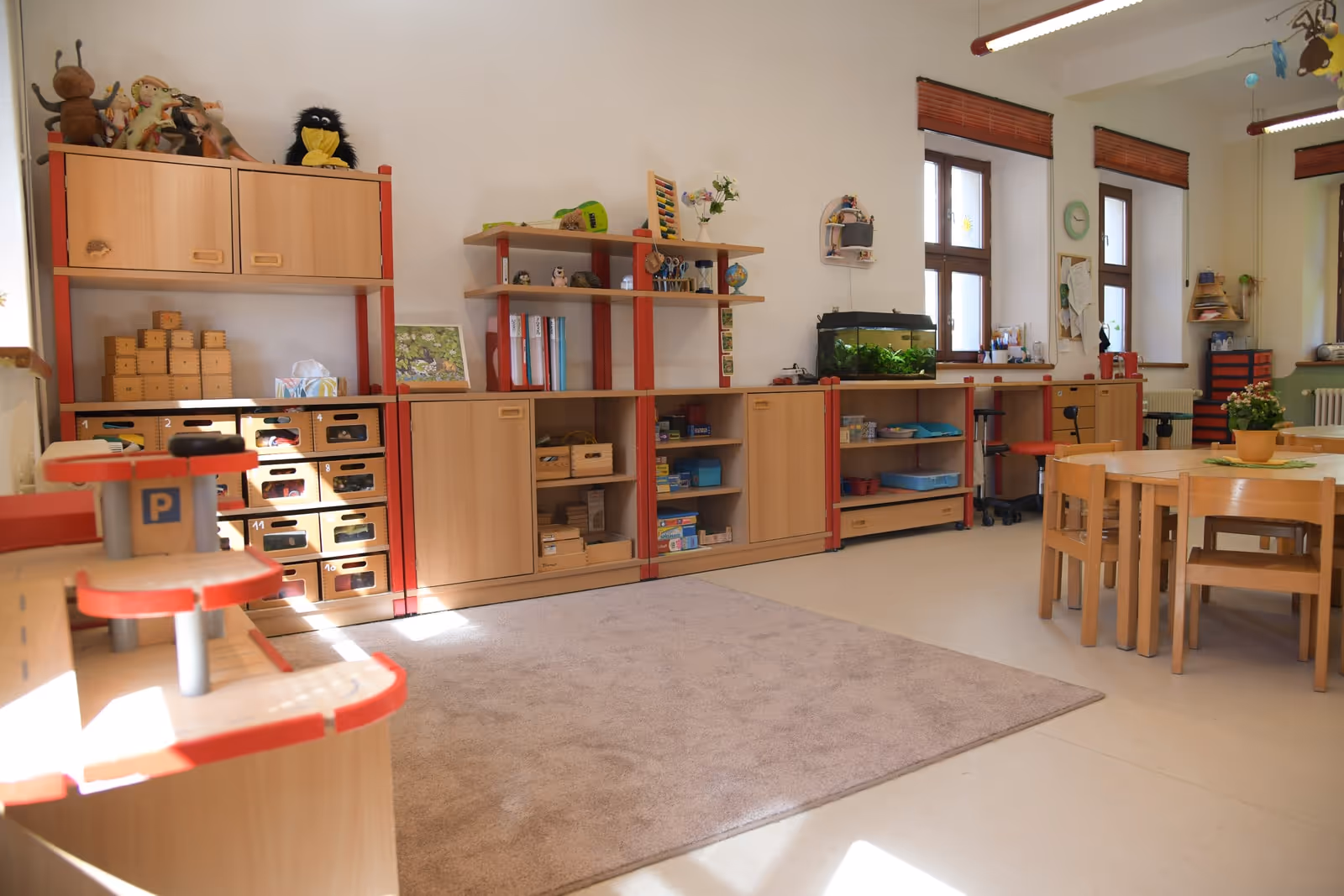 Gruppenraum mit Holzregalen voller Spielmaterial, Kinderküche und Tischen am Fenster.
