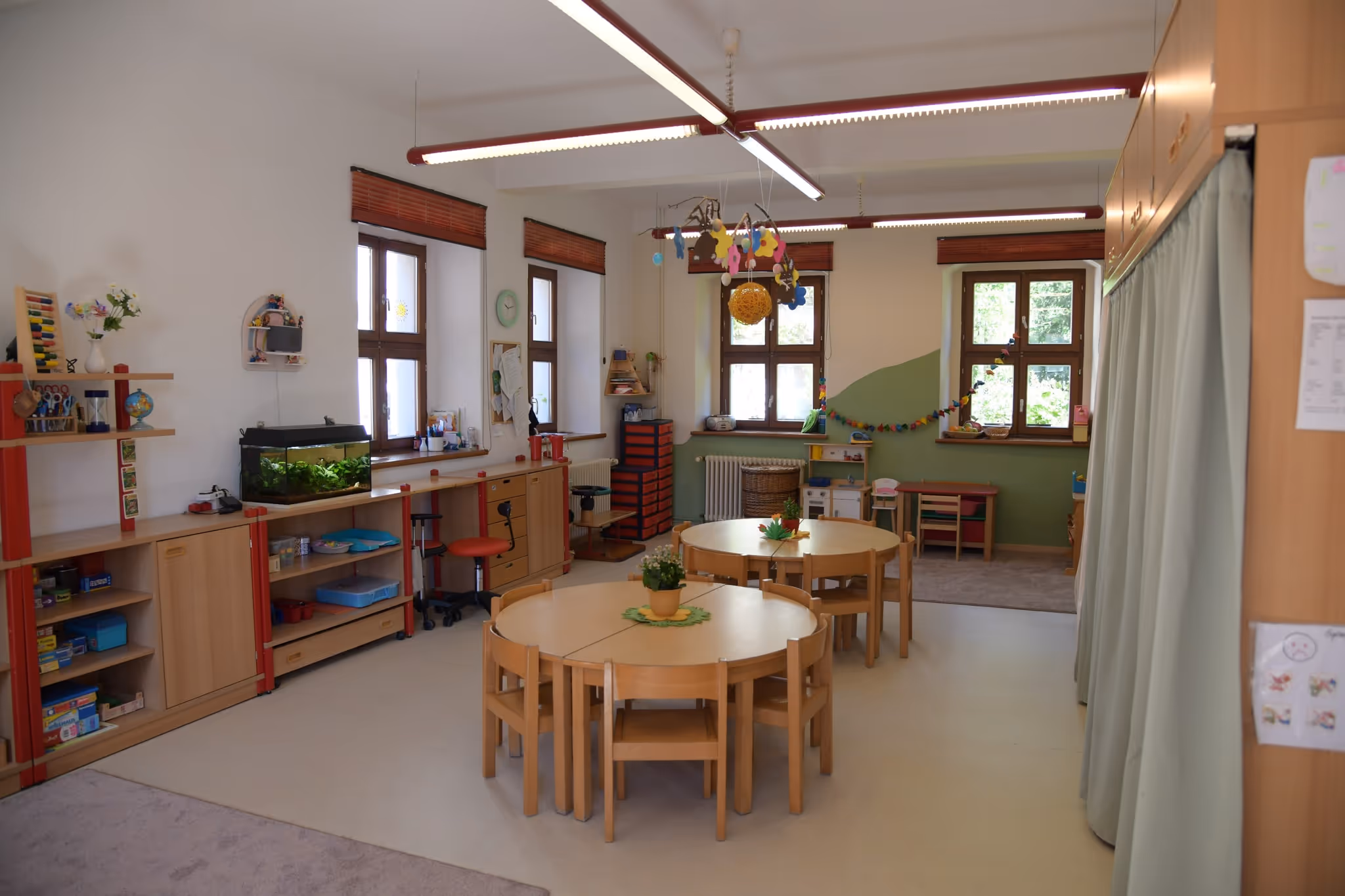 Heller Gruppenraum mit runden Holztischen, Kinderstühlen und niedrigen Regalen vor großen Fenstern.