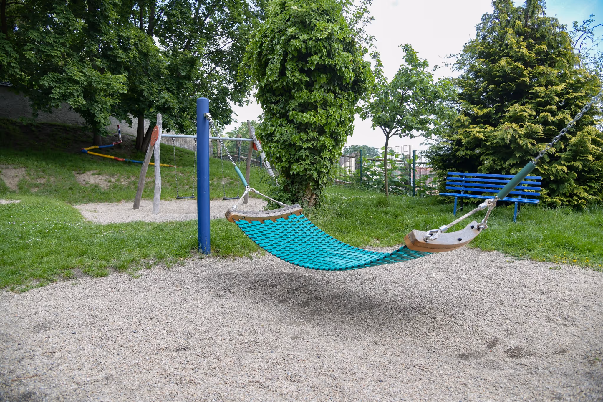 Türkise Hängematte zwischen Pfosten auf einem sandigen Spielbereich mit Bäumen und Sitzbank.