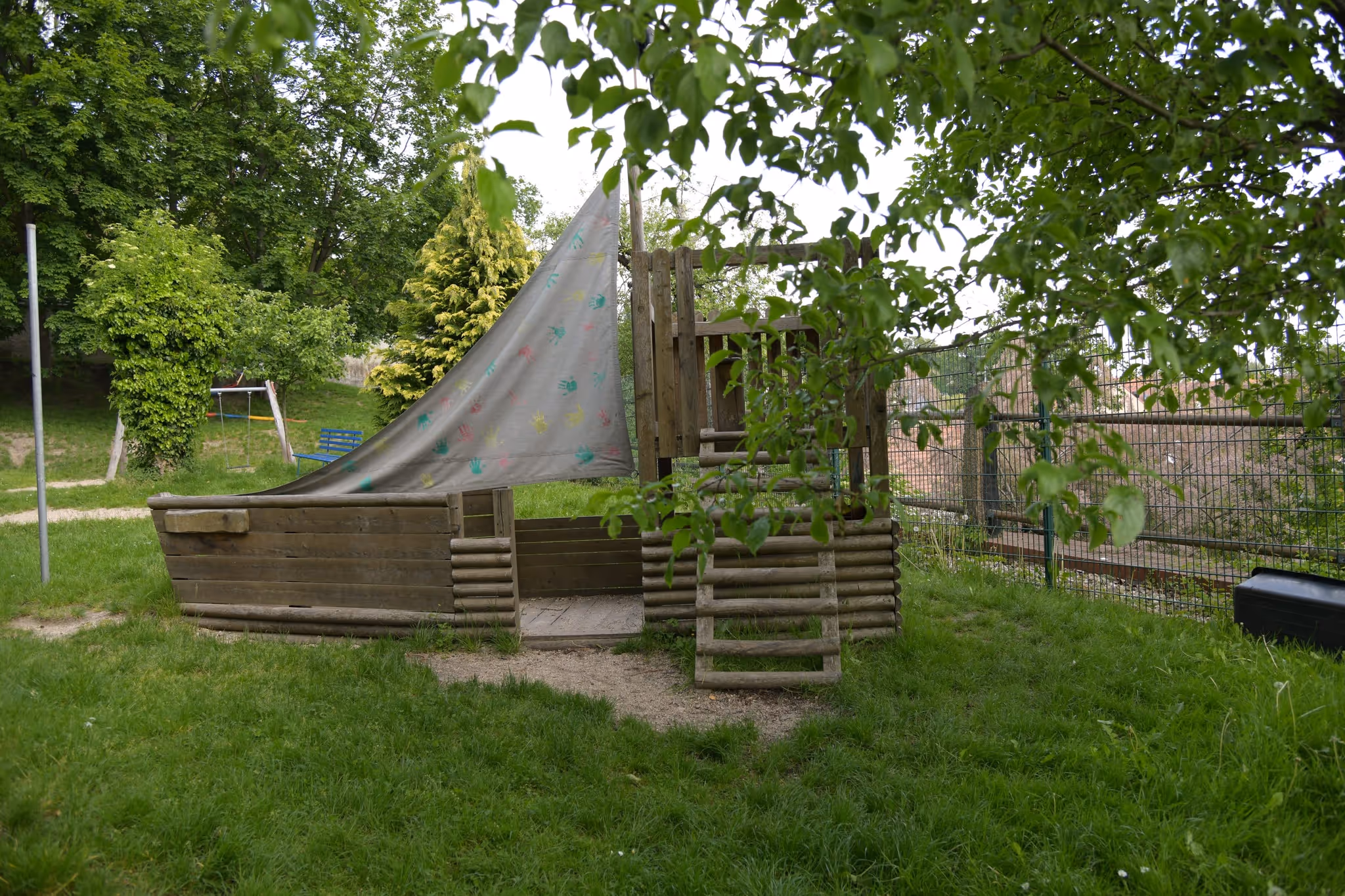 Hölzernes Spielschiff mit Sonnensegel auf einer Wiese im Außengelände.