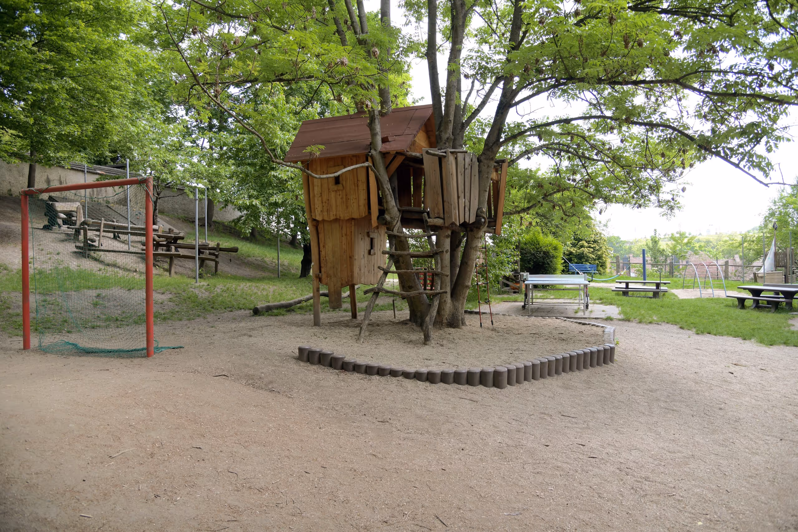 Hölzernes Spielhaus auf Stelzen zwischen Bäumen, umgeben von einem sandigen Spielbereich.