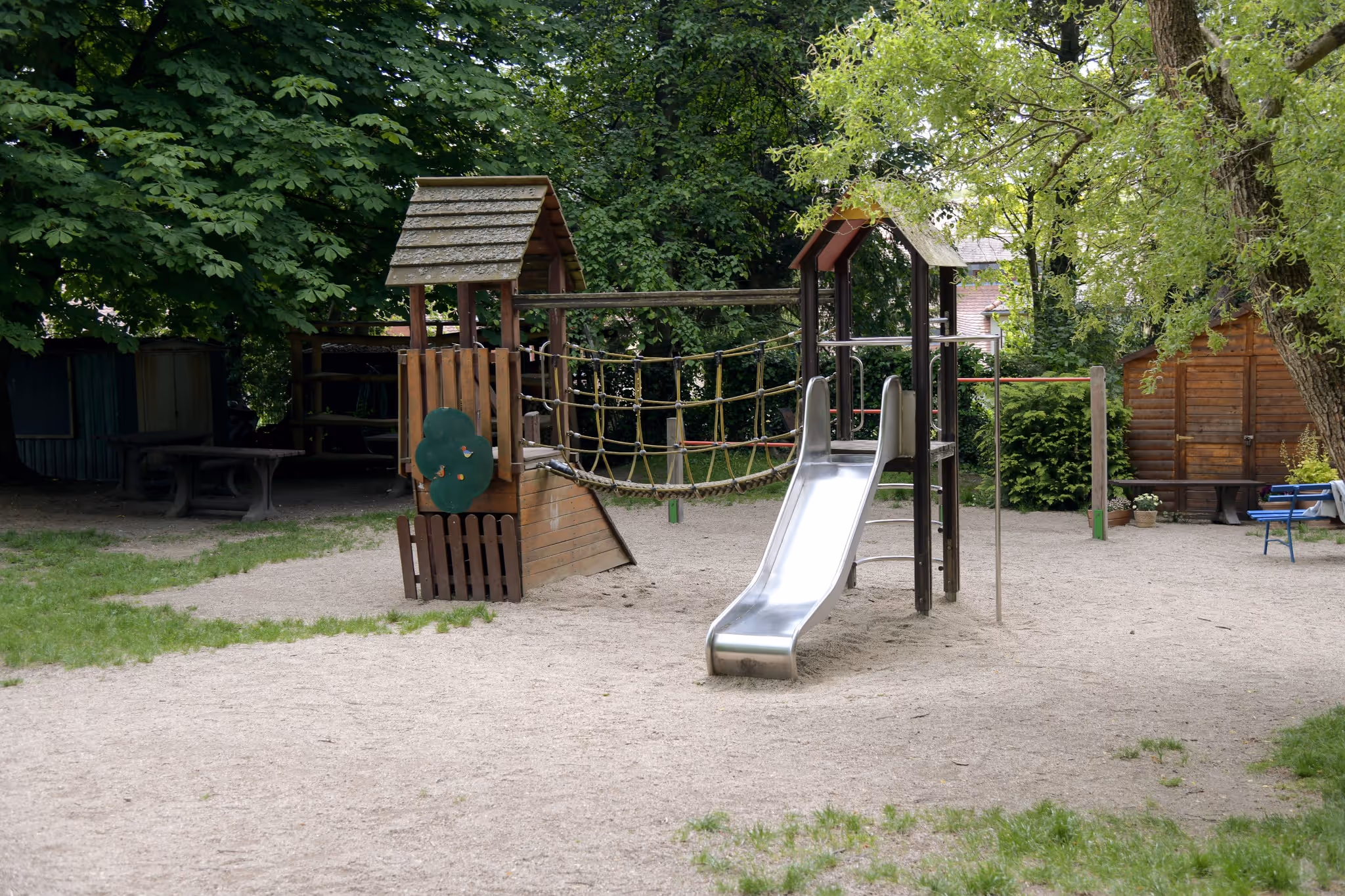 Hölzernes Klettergerüst mit Metallrutsche, Kletternetz und Schaukel auf einem Sandspielplatz.