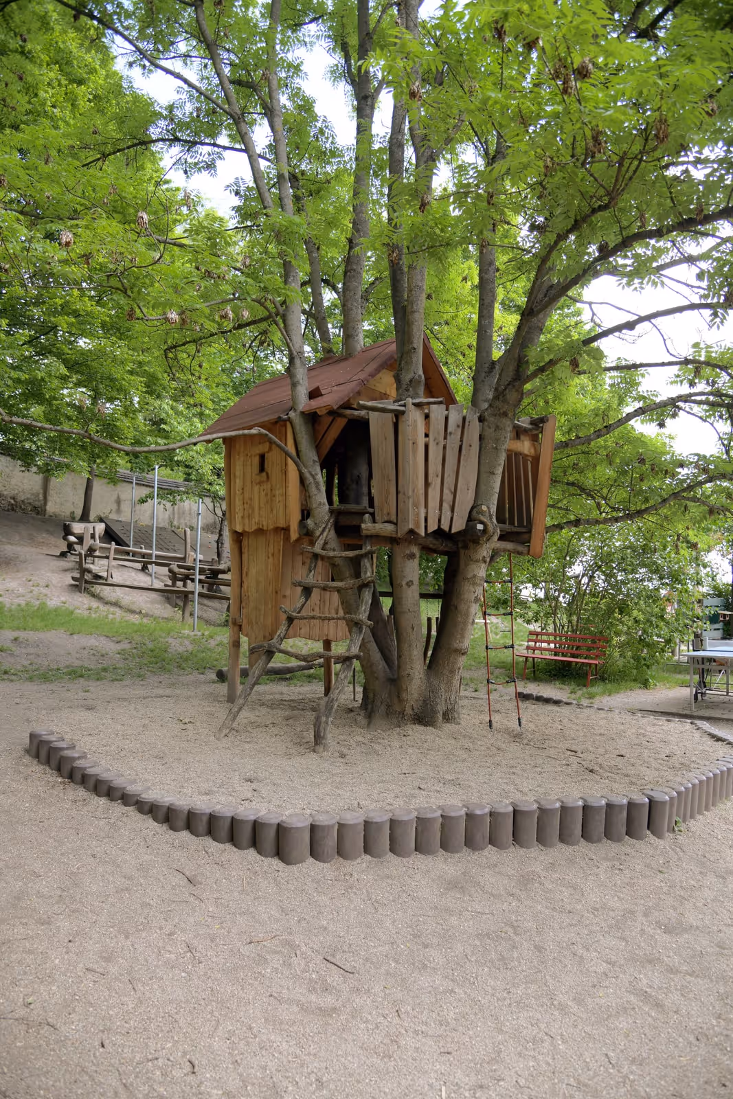 Spielhaus aus Holz auf Stelzen mit Leiter und Balkon, umgeben von Bäumen.