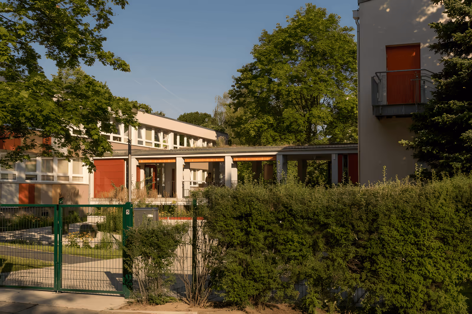 Gebäude mit großem grünem Tor, Hecke und Bäumen unter klarem blauem Himmel.