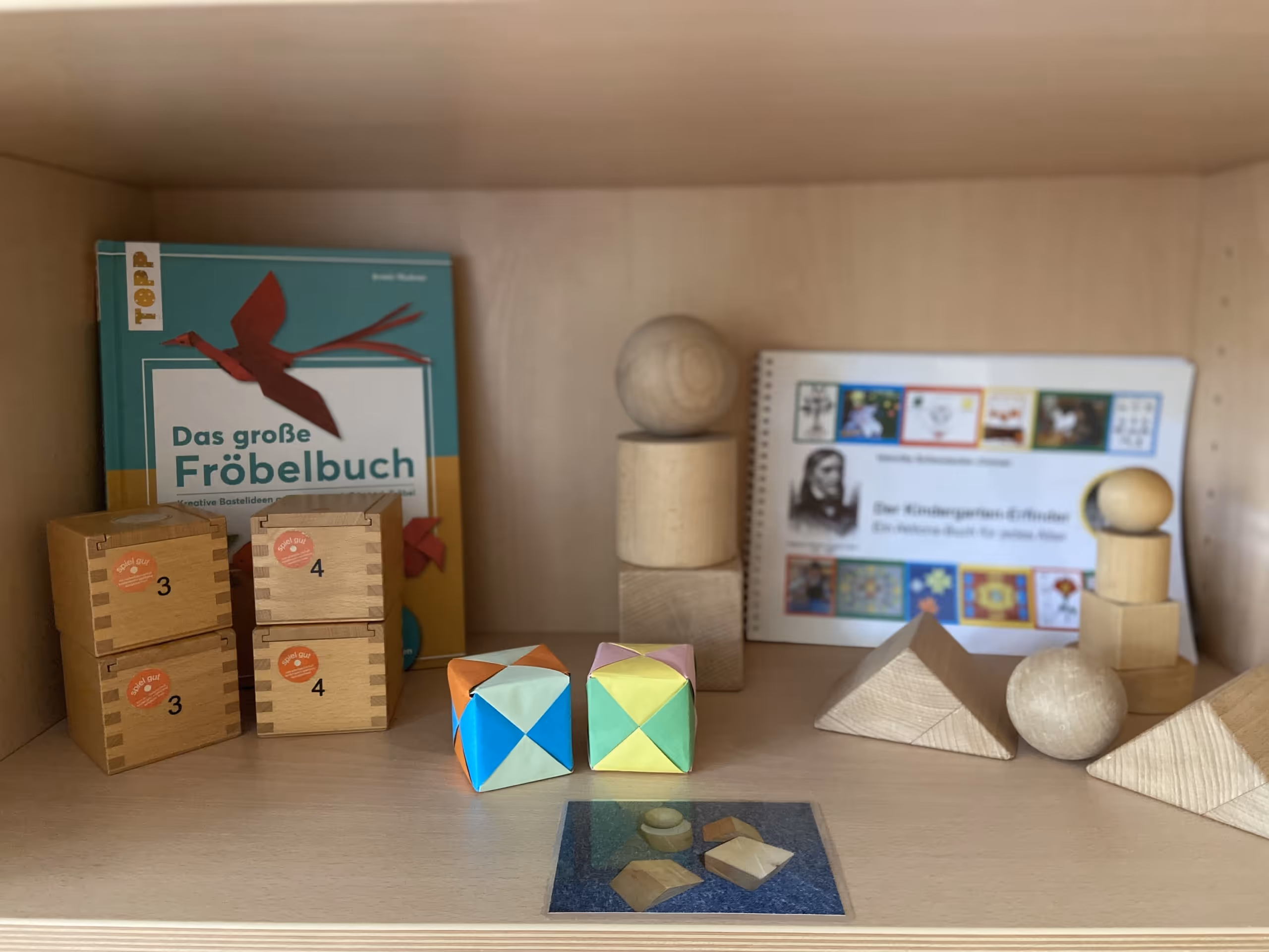 Fröbelgaben im Regal: Holzwürfel, Kugeln und Farbtäfelchen neben dem Buch „Das große Fröbelbuch“.
