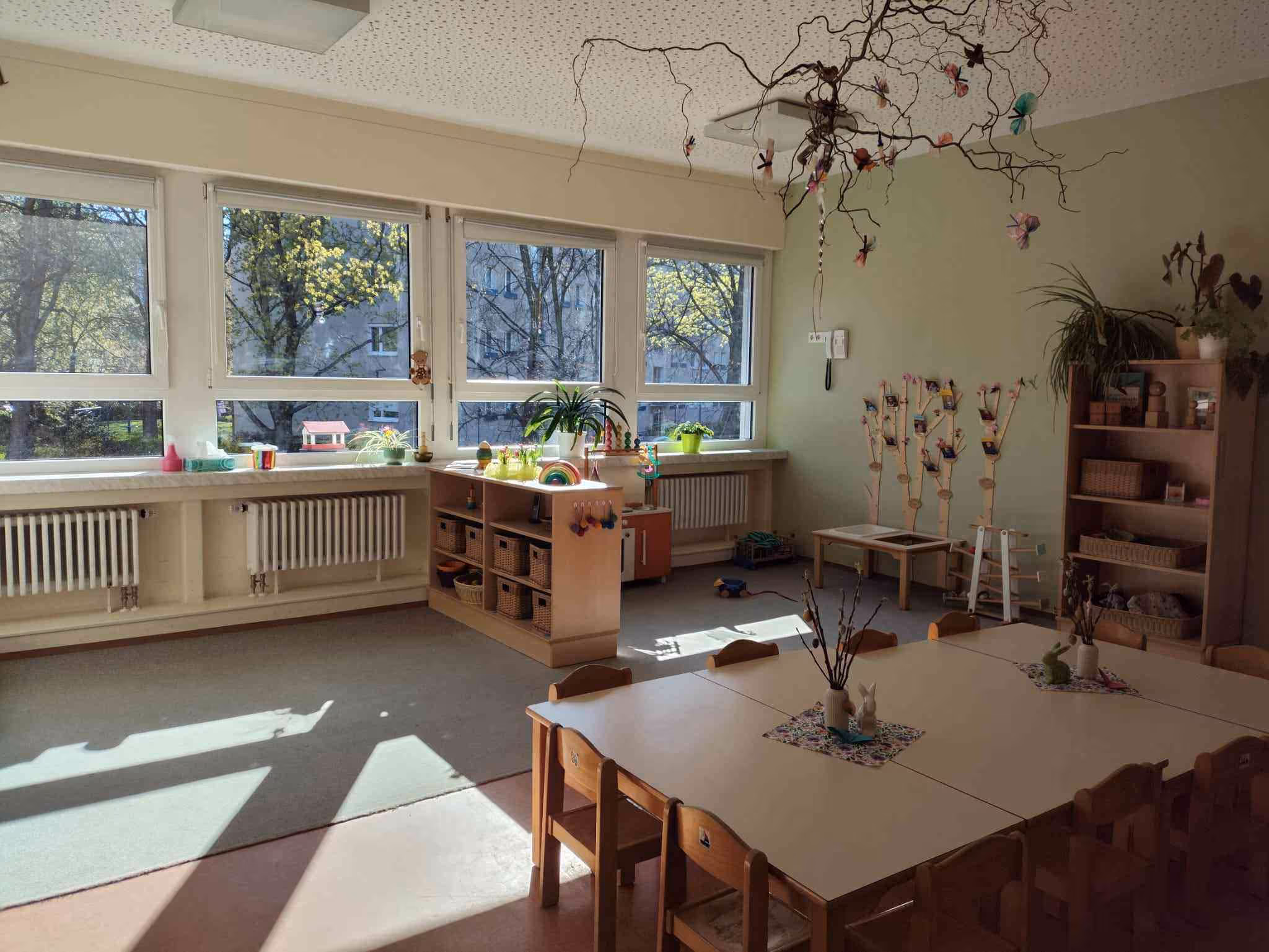 Gruppenraum „Spatzen“ mit Kindertischen, Materialregalen und Fensterfront zum Garten.