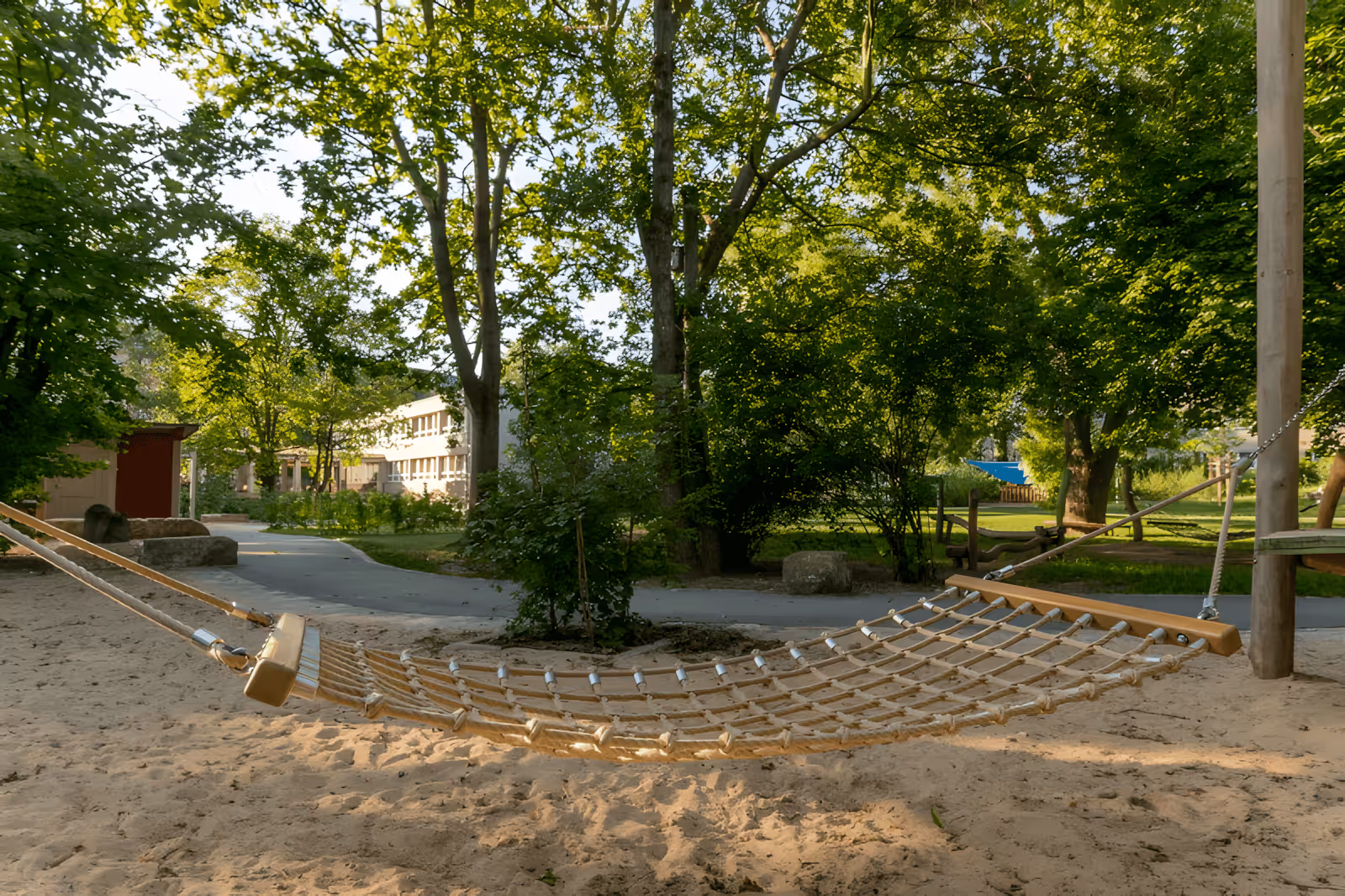 Geflochtene Hängematte aus Seilen über Sand in einem sonnigen Park mit Bäumen.