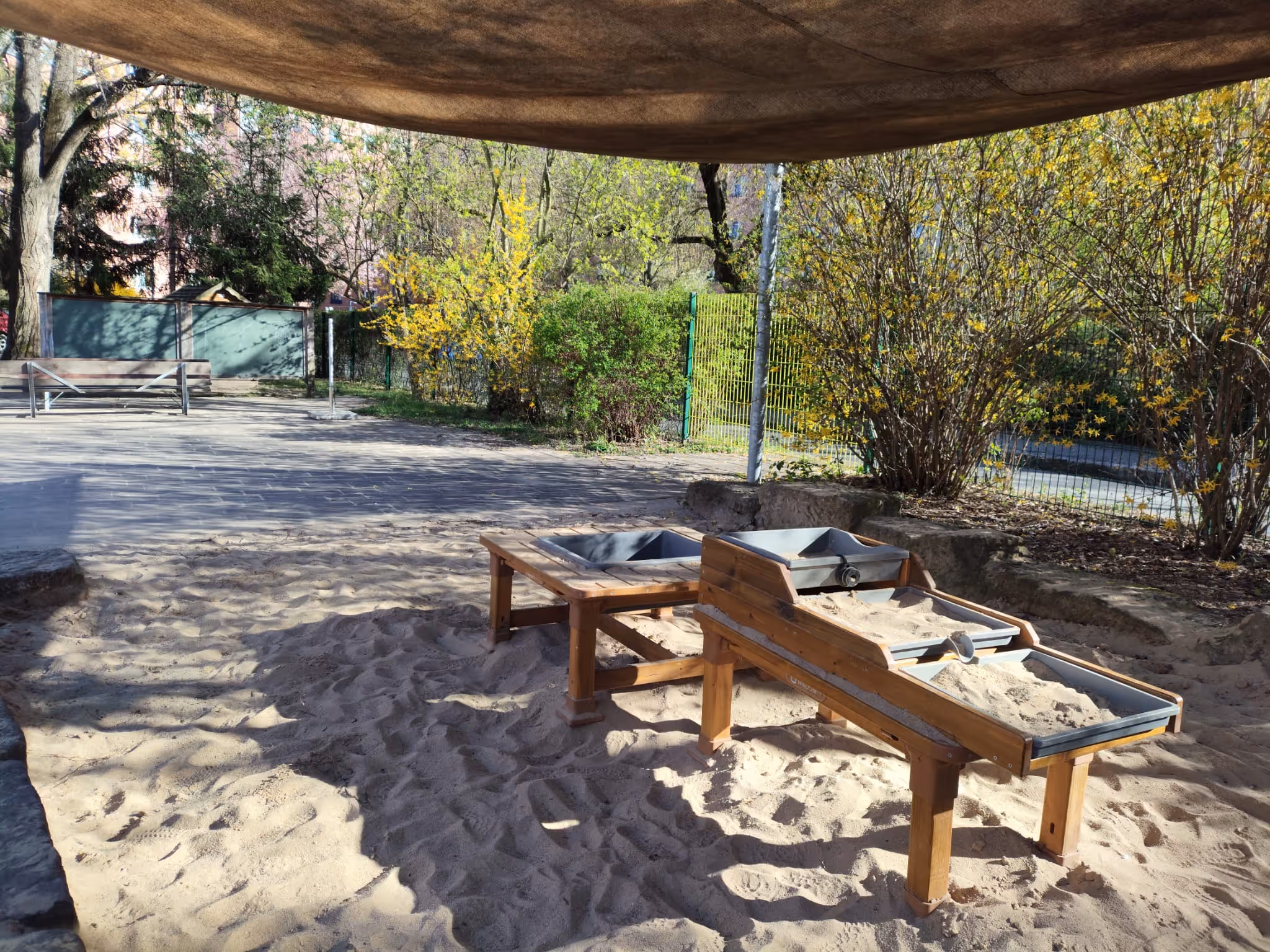 Sandkasten mit Holzrahmen und Sand im Vordergrund, Bäume und Bänke im sonnigen Park.