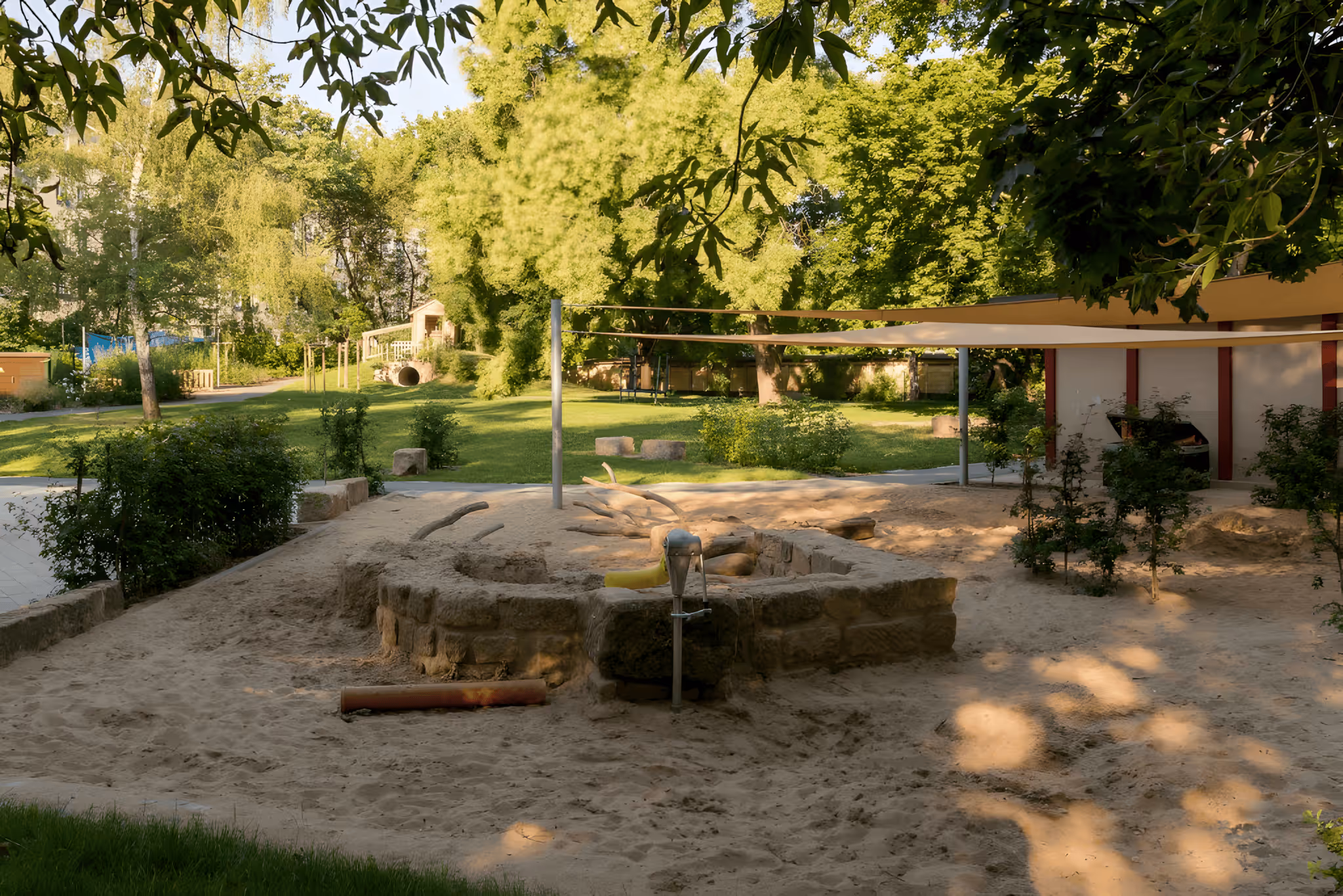 Sandkasten mit Steinumrandung und Wasserpumpe in einem sonnigen, grünen Kinderspielplatz.