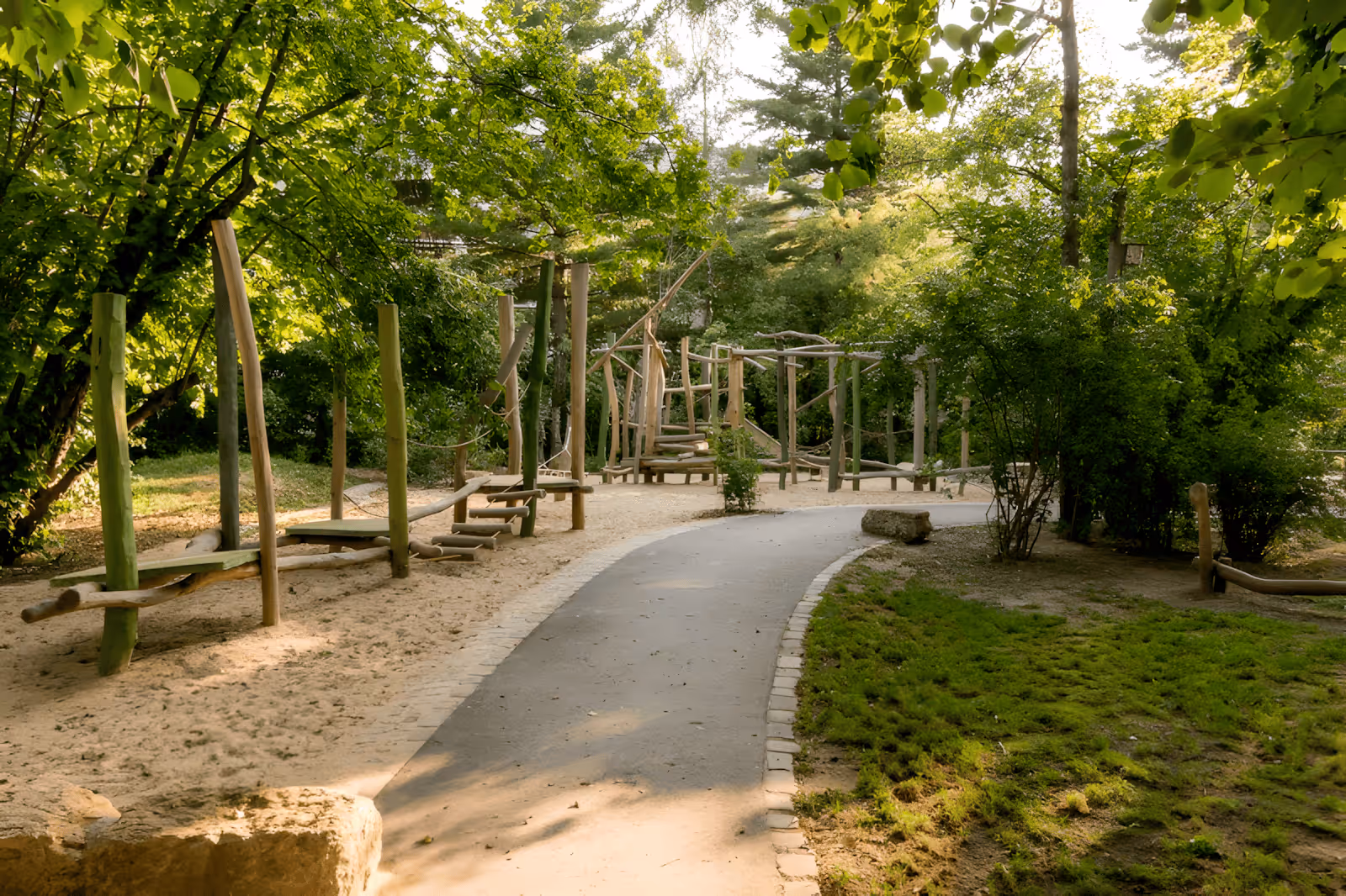 Waldspielplatz mit Holz-Klettergerüst und gepflastertem Weg umgeben von grünen Bäumen.